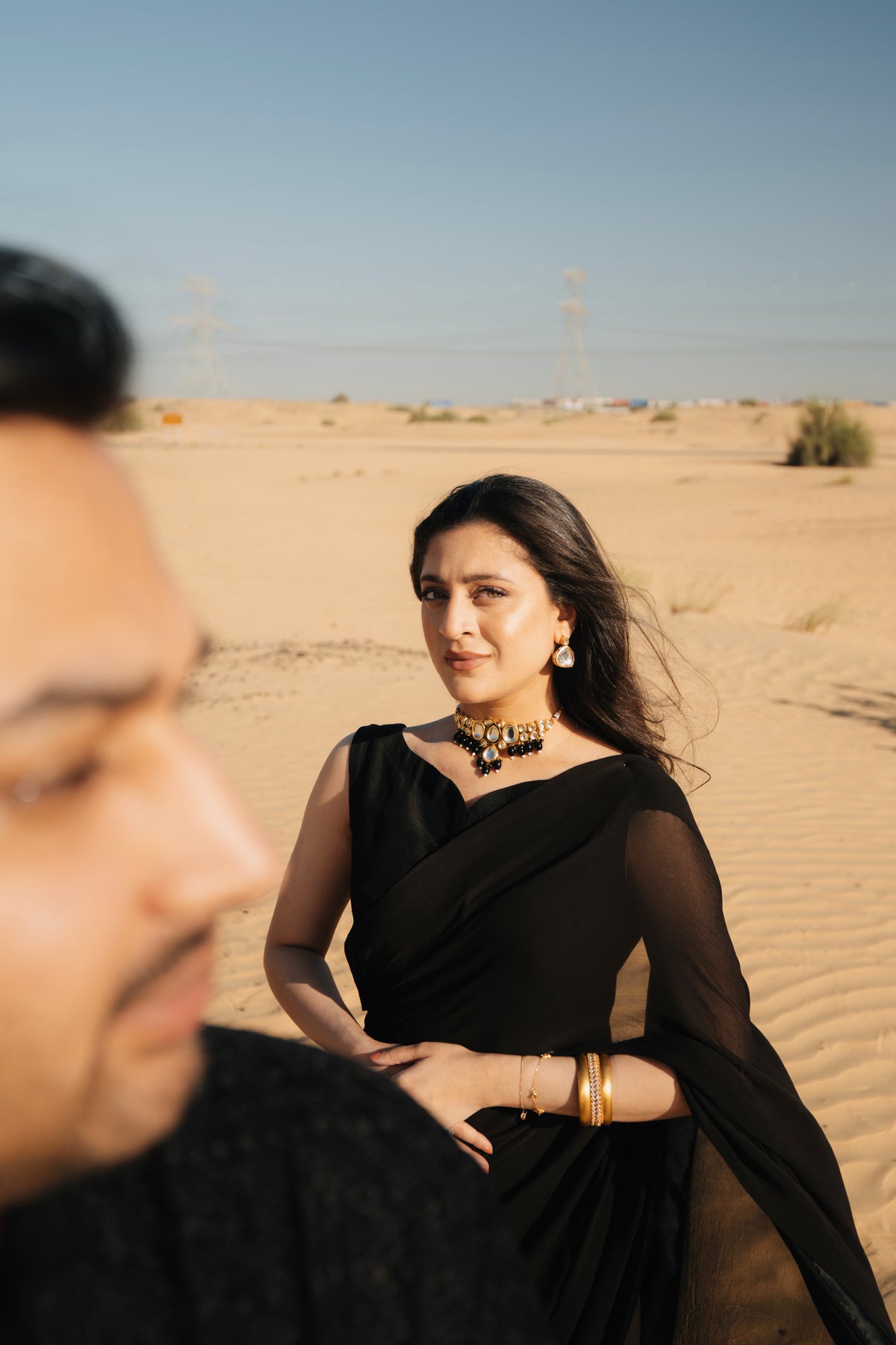 A woman wearing a black sari and gold jewelry stands in a sandy desert, with a man partially visible in the foreground.